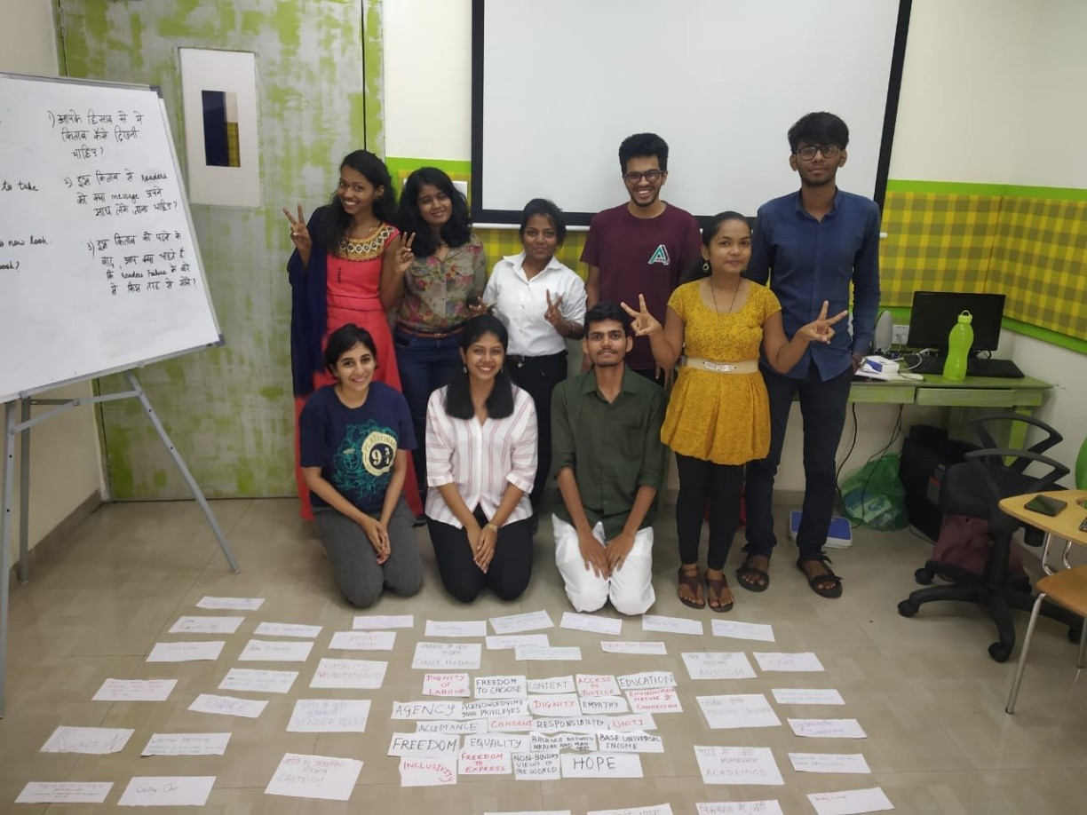 Group photo in a classroom: four participants standing in the back row, three kneeling in front. On the floor in front of them, cards spell out words like FREEDOM, AGENCY, EDUCATION, INCLUSION, JUSTICE, HOPE.