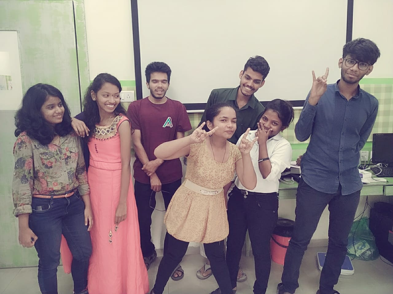 Seven young participants posing playfully together in a classroom - peace signs, smiles, leaning in close.